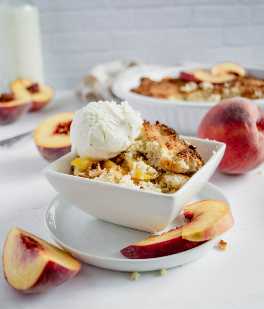 peach cobbler in a white bowl surrounded by peaches and topped with ice cream