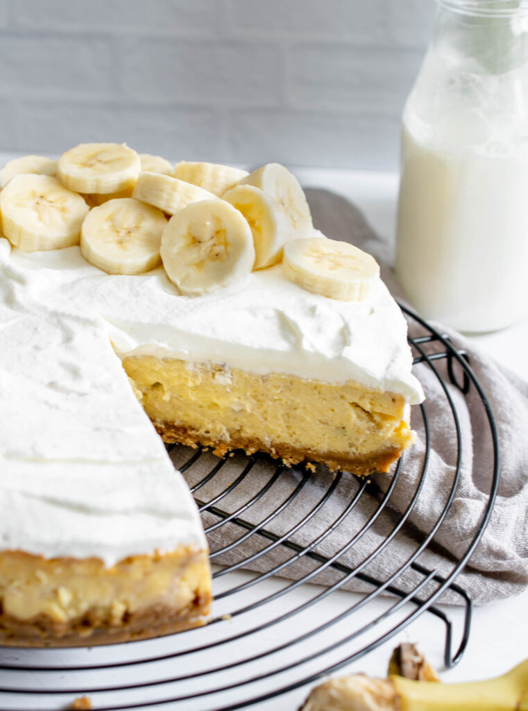 banana pudding cheesecake on a wire cooling rack with one slice missing. Topped with whipped cream and sliced bananas