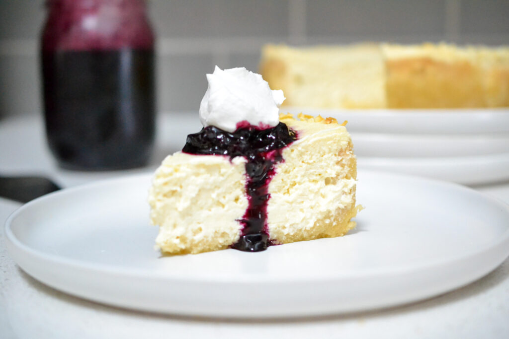slice of cheesecake on a white plate topped with blueberry jam and whipped cream
