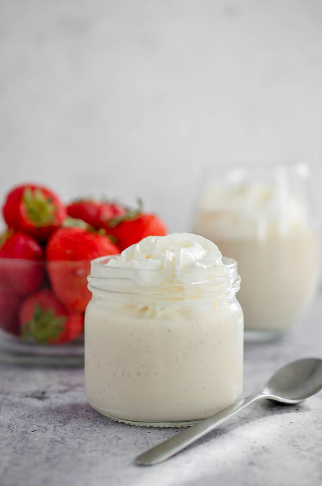 Vegan Keto Vanilla Pudding in a small glass jar topped with whipped cream, bowl of strawberries in the background